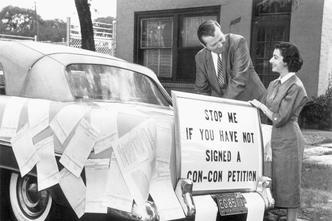A black and white photo. There's a sign on the back of a car that says, "Stop me if you have not signed a con-con petition." 